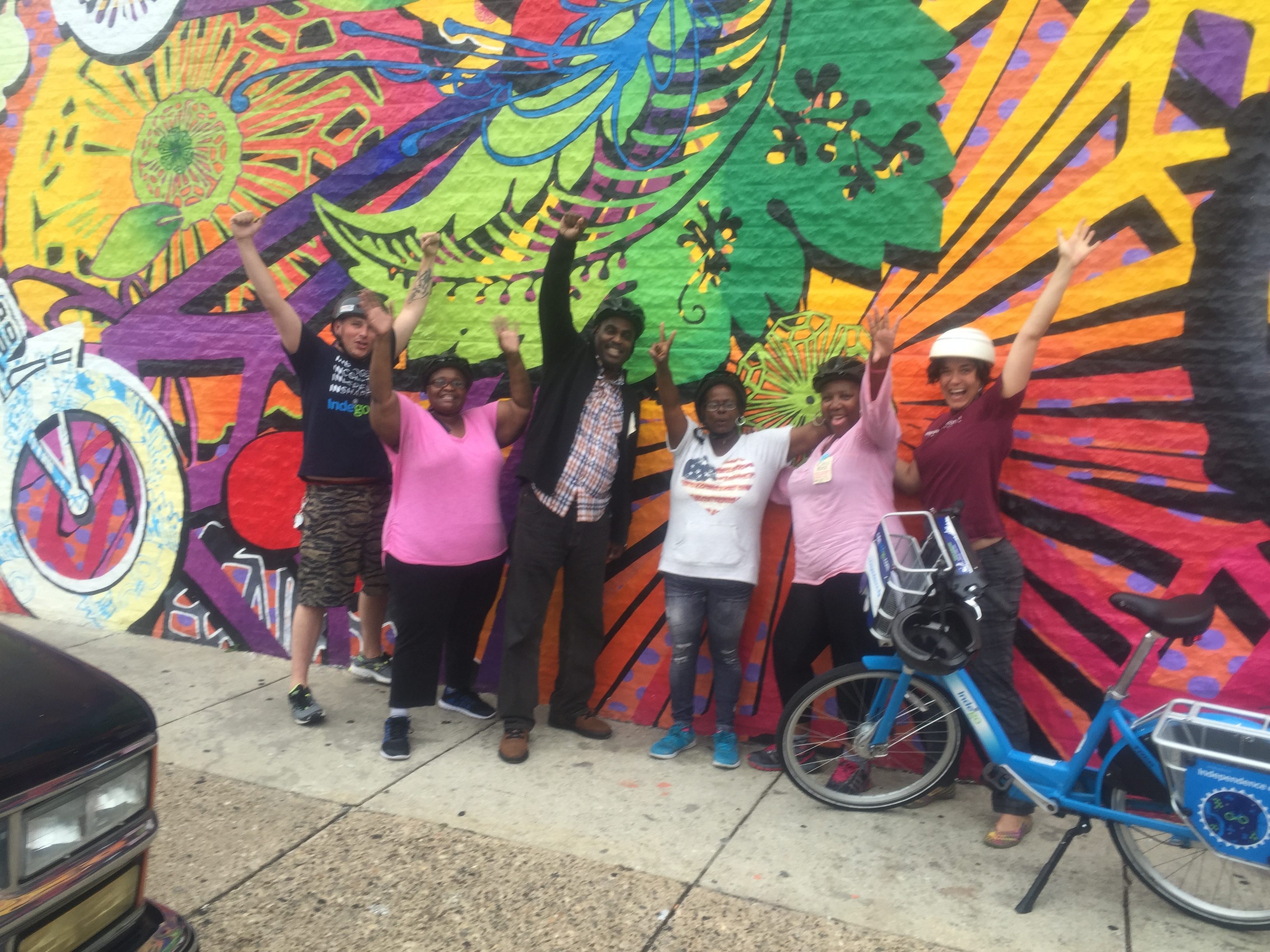 A group of participants and staff pose smiling in front of a colorful mural. They are wearing bike helmets and stand next to a bike,